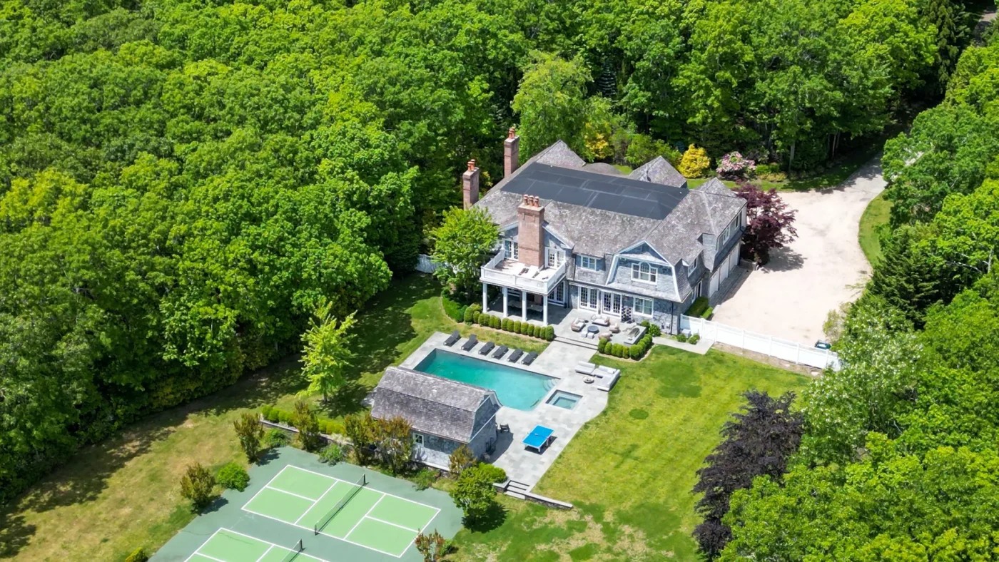 Aerial view of the estate at 486 Middle Line Hwy in Bridgehampton, featuring a large house, pool, and tennis court surrounded by lush greenery.