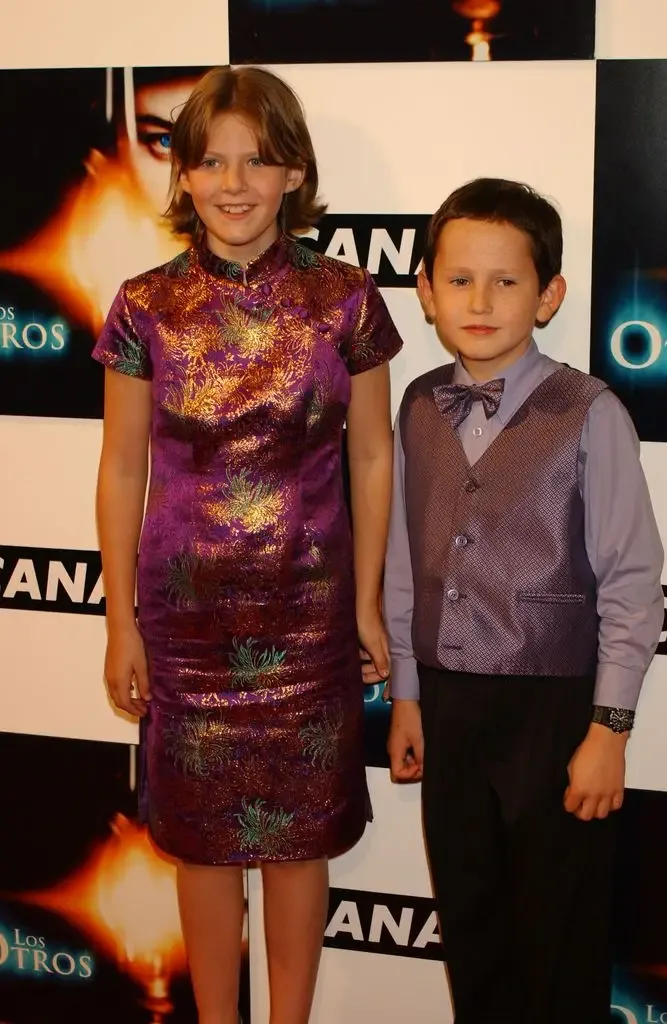 Alakina Mann in a purple silk dress and James Bentley in a bow tie and waistcoat pose together in front of a 'Los Otros' promotional backdrop.