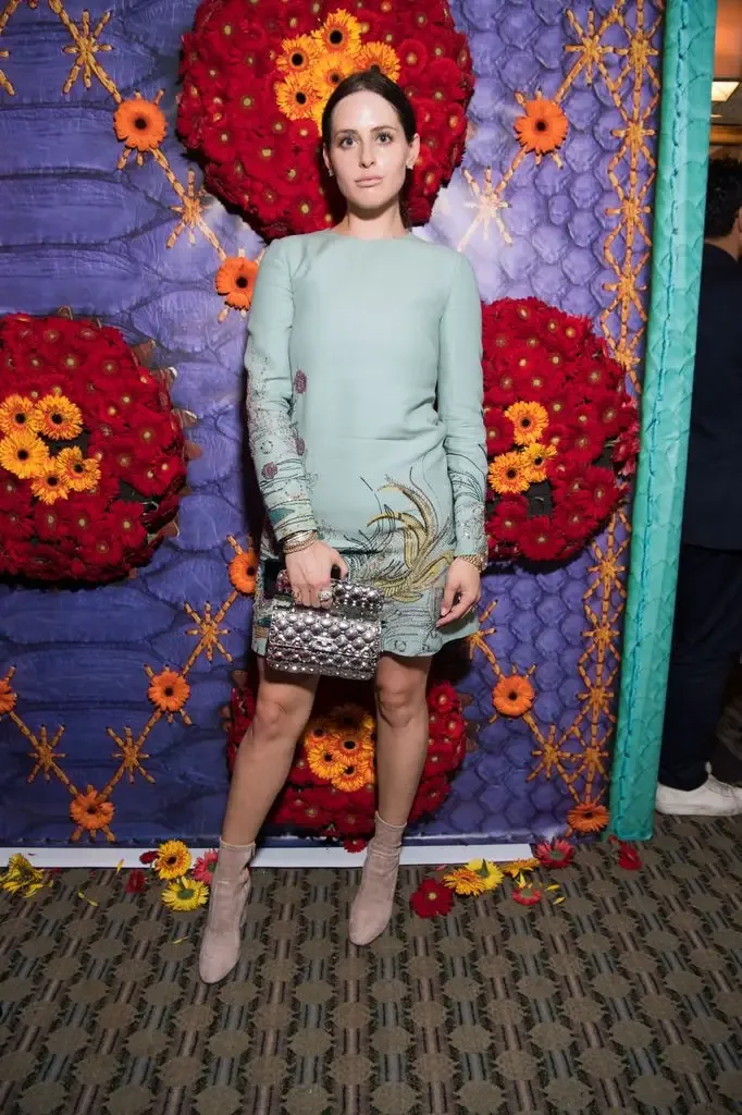 Alexa Dell, wearing a teal dress and nude boots, stands before a colorful floral backdrop