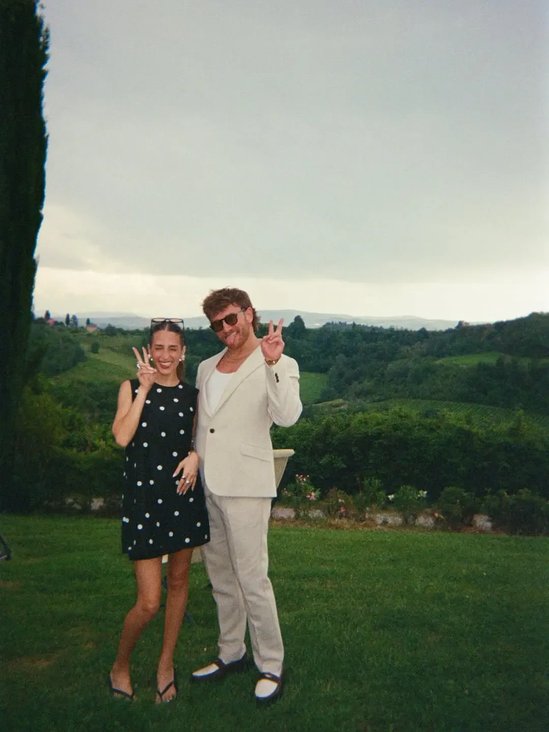 Amanda Batula and West Wilson posing outdoors, both flashing peace signs against a scenic rural landscape backdrop.