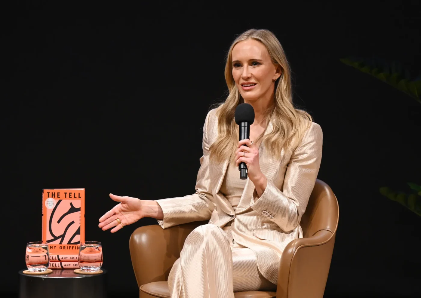 Amy Griffin speaks onstage holding a microphone while seated in a chair next to a table with a copy of her book,