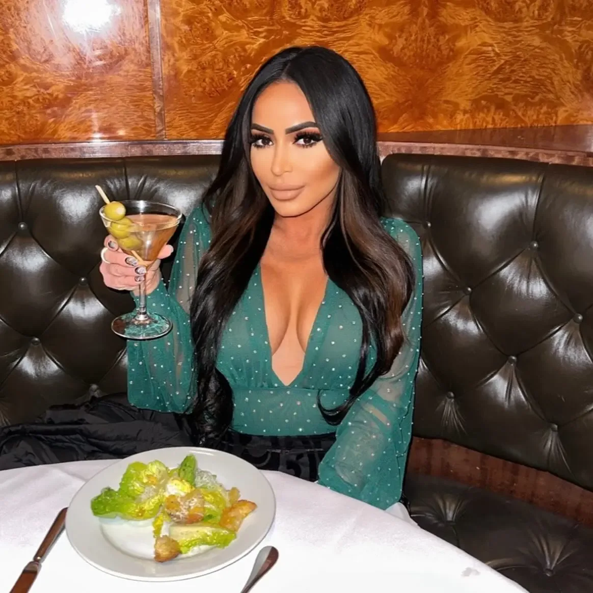 Angelina Pivarnick sitting in a booth holding a martini with a salad on the table in front of her.