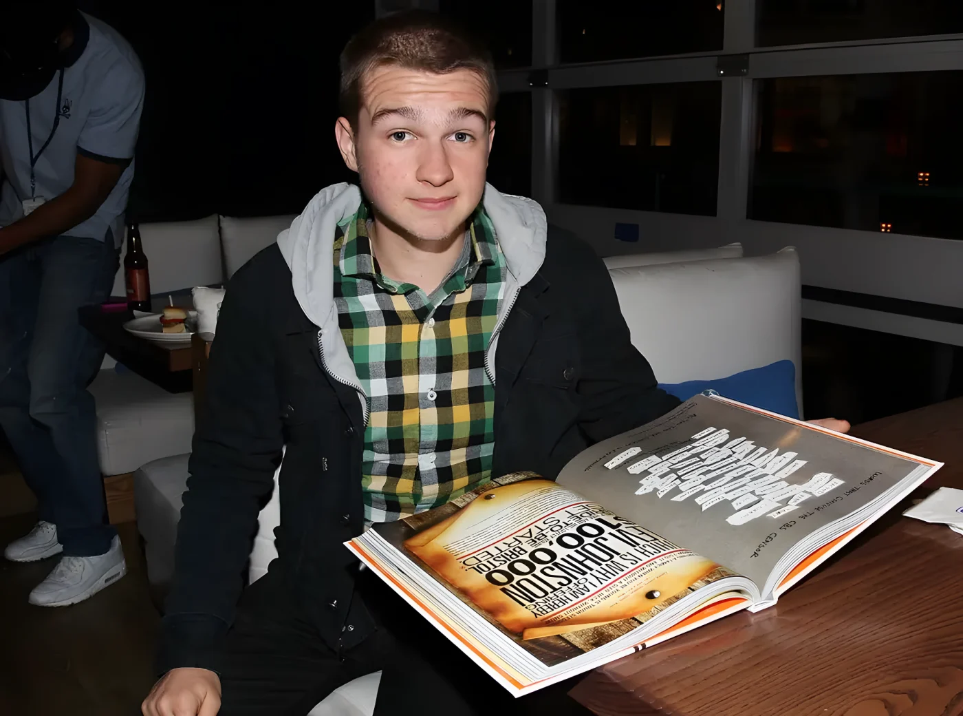 Angus T. Jones sits holding an open book featuring Chuck Lorre vanity card text, smiling at the camera.