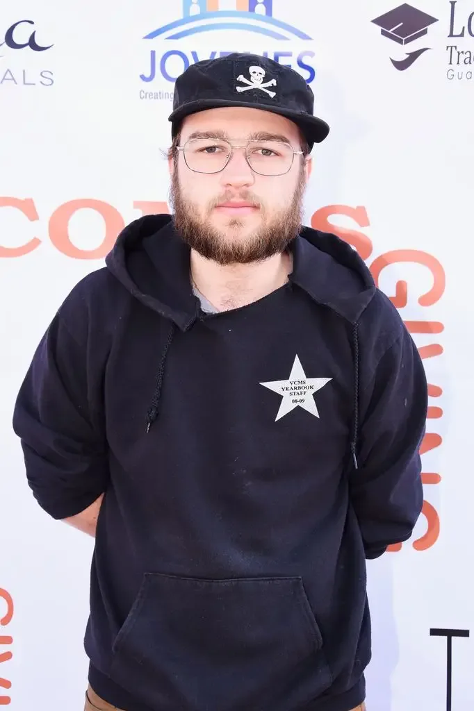 Angus T. Jones, wearing a cap, glasses and a black hoodie, poses for photographers at an event step and repeat.