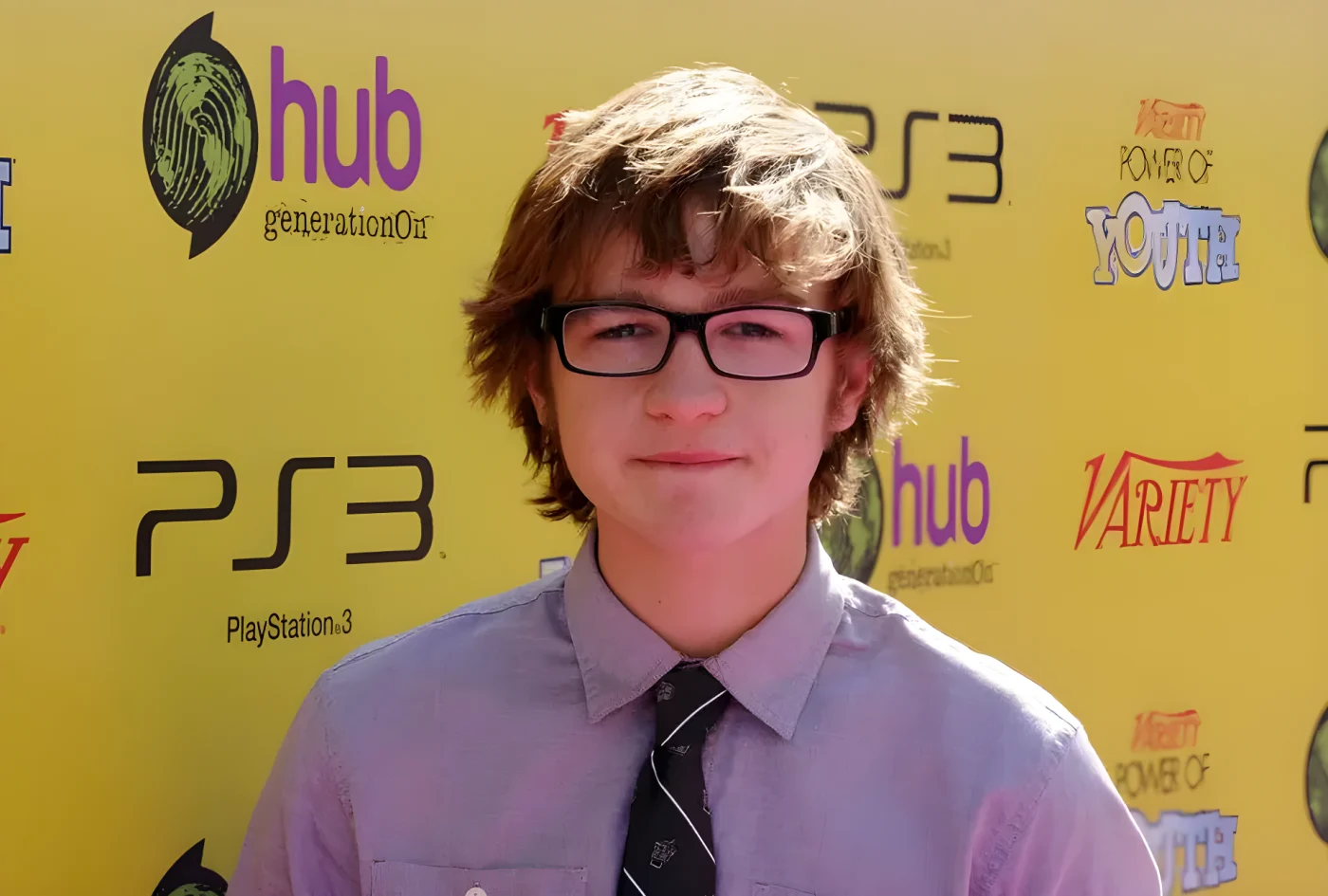 Angus T. Jones, wearing glasses and a tie, poses on the red carpet in front of a yellow step-and-repeat backdrop.