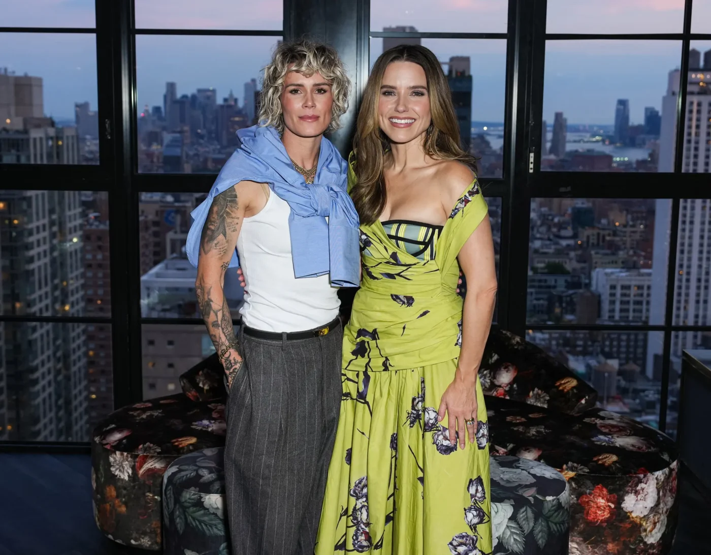 Ashlyn Harris and Sophia Bush pose together in front of a window with a city skyline view.