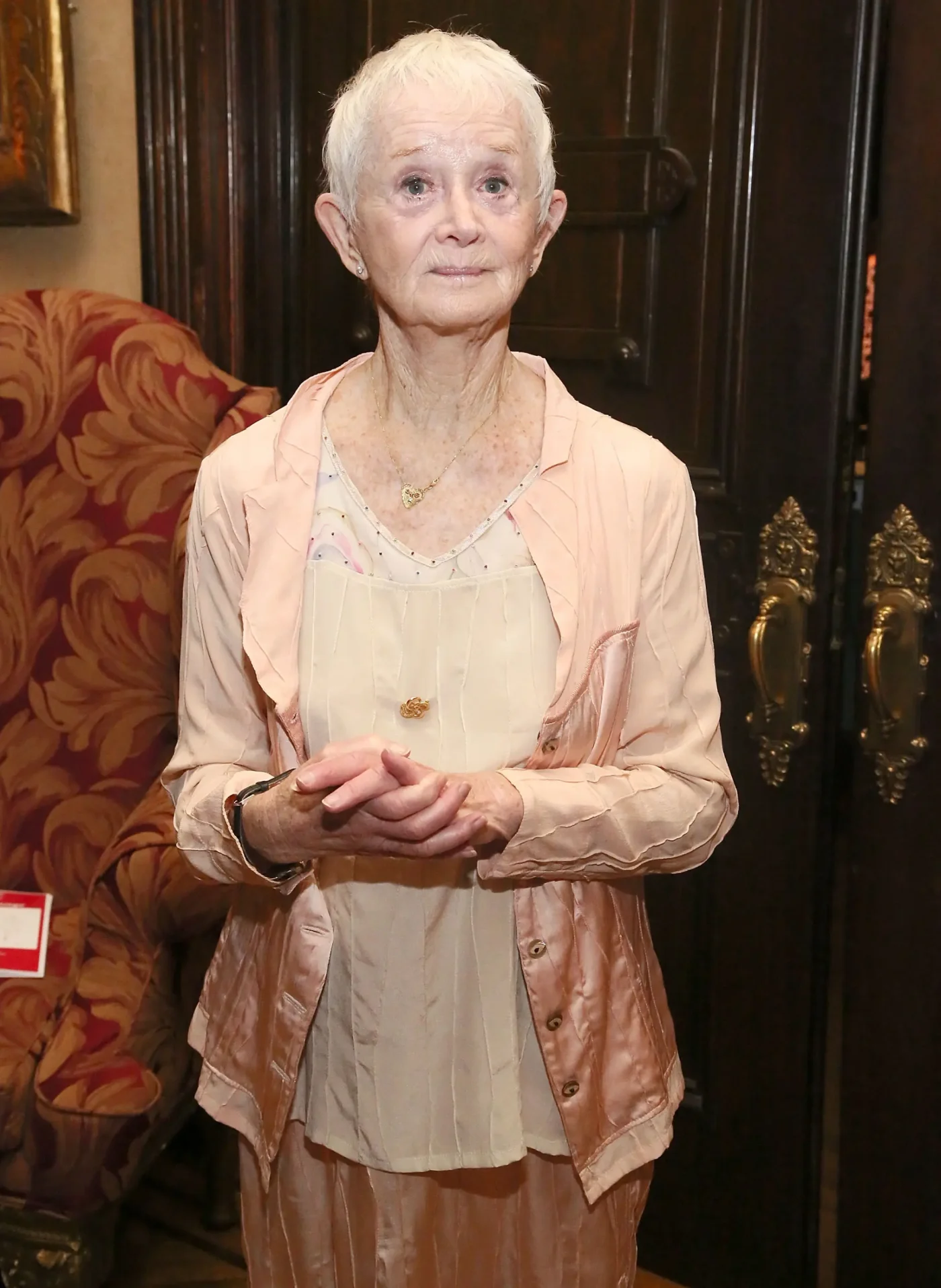 Barbara Barrie standing indoors and wearing a light pink jacket.