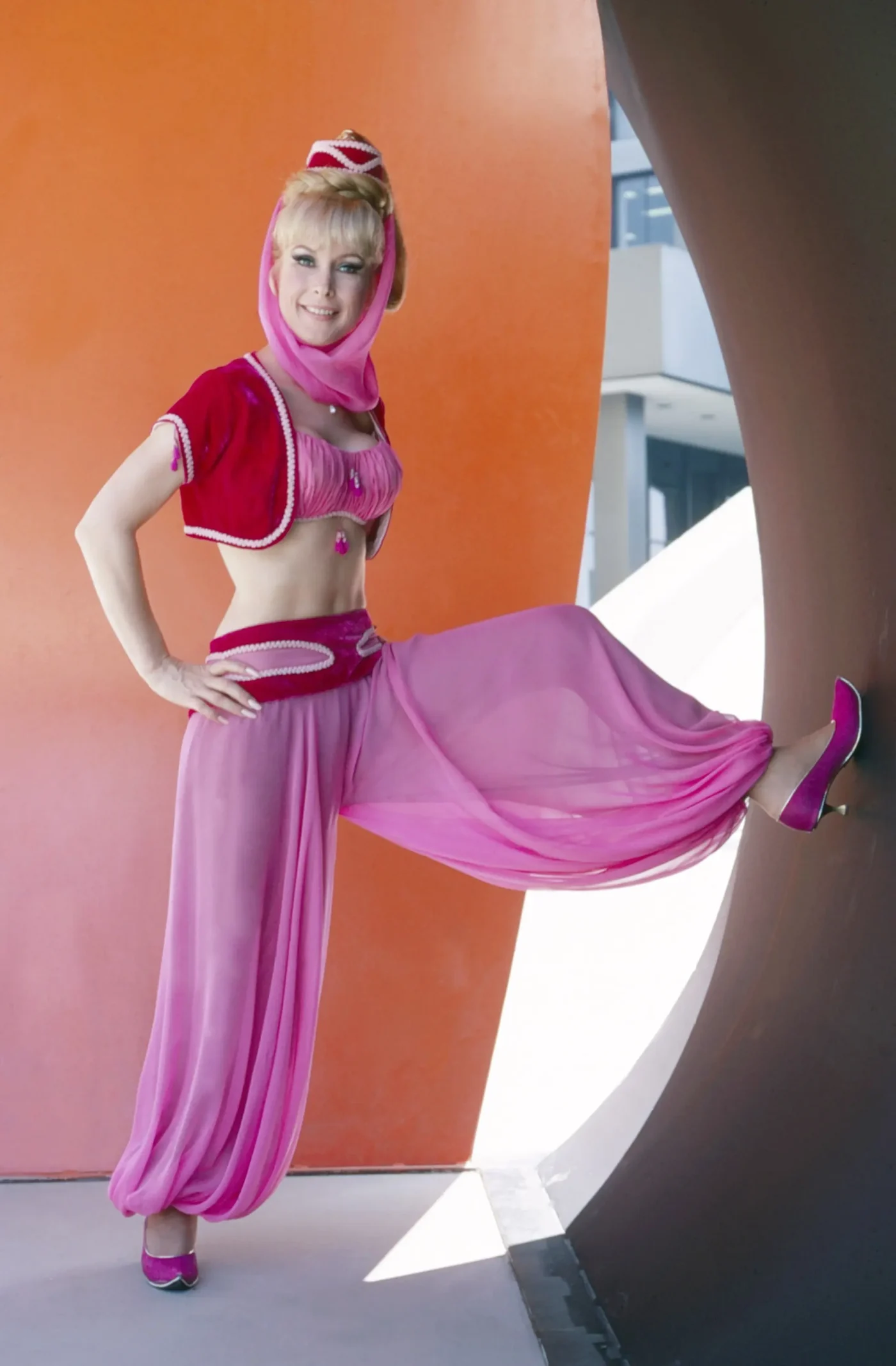 Barbara Eden as Jeannie, wearing a pink harem costume with a matching veil and vest, posing in front of an abstract orange and brown structure.