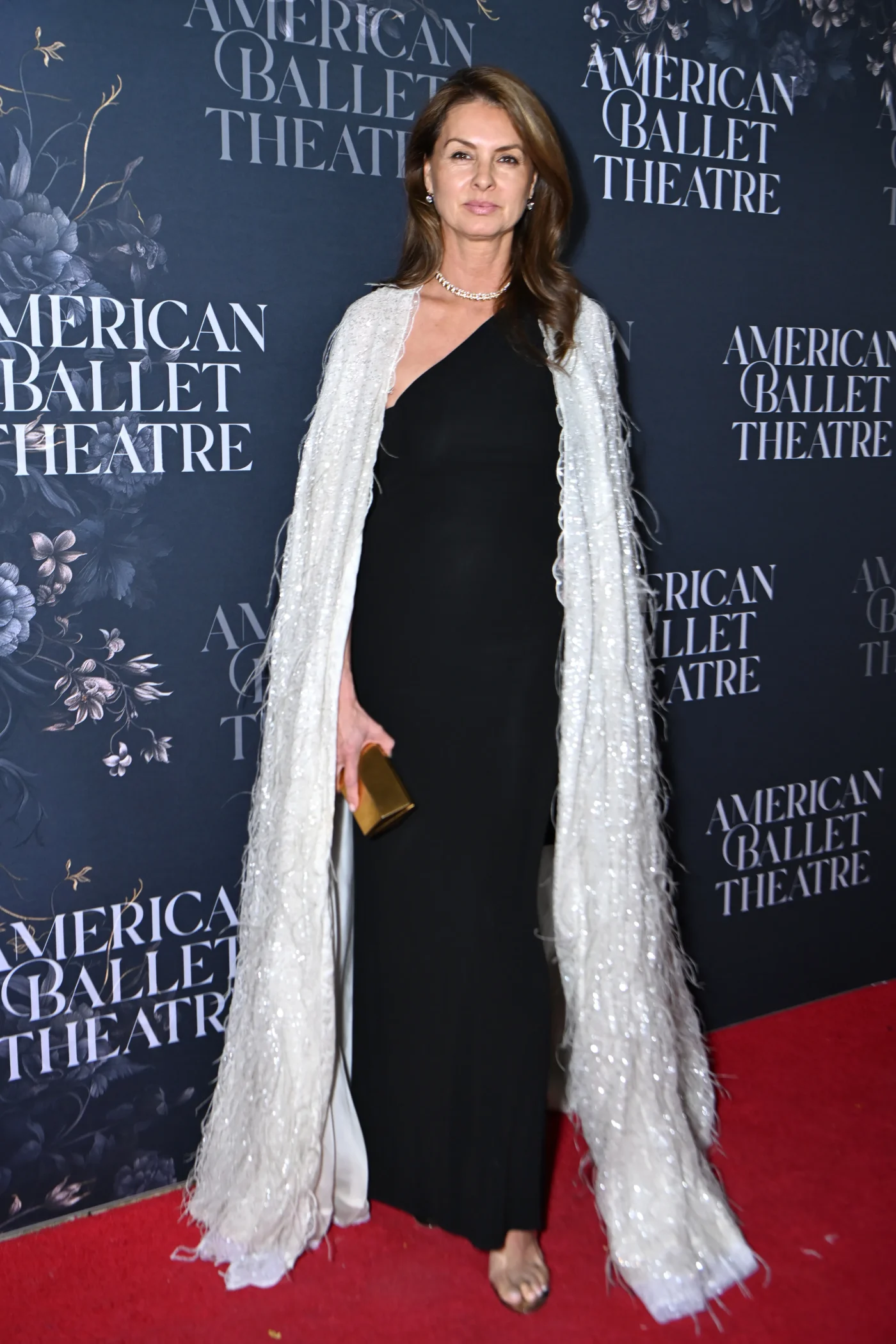 Barbara Siegler poses on the red carpet at the American Ballet Theatre Fall Gala, wearing a black one-shoulder gown paired with a long, sparkly white cape and holding a gold clutch.