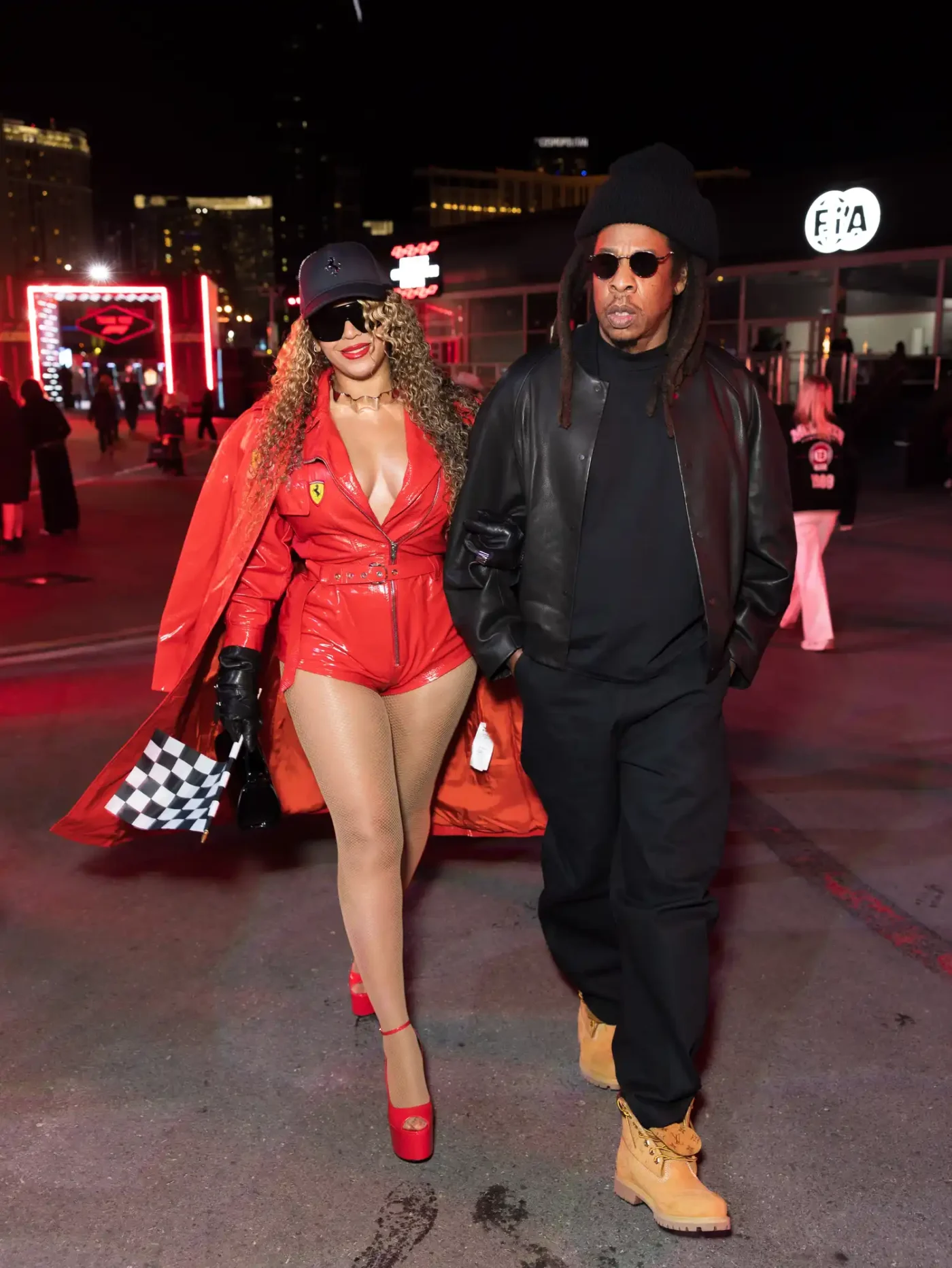 Beyoncé and Jay-Z walk side by side at the Las Vegas Strip Circuit. Beyoncé is wearing a plunging red leather jumpsuit with micro shorts, a matching jacket, and strappy platform heels. Jay-Z is wearing an all-black ensemble, including a leather jacket, and tan work boots.
