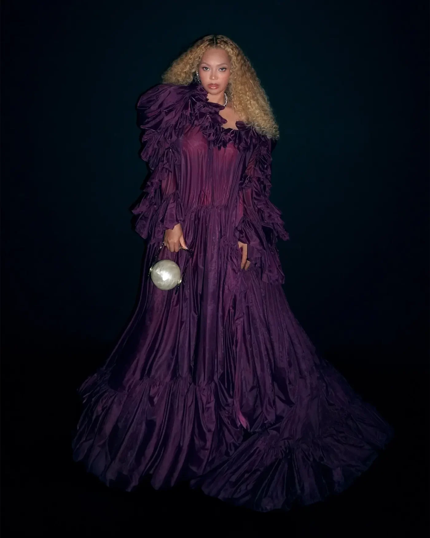 Beyonce wearing a ruffled purple dress and holding a sphere-shaped handbag against a dark background.