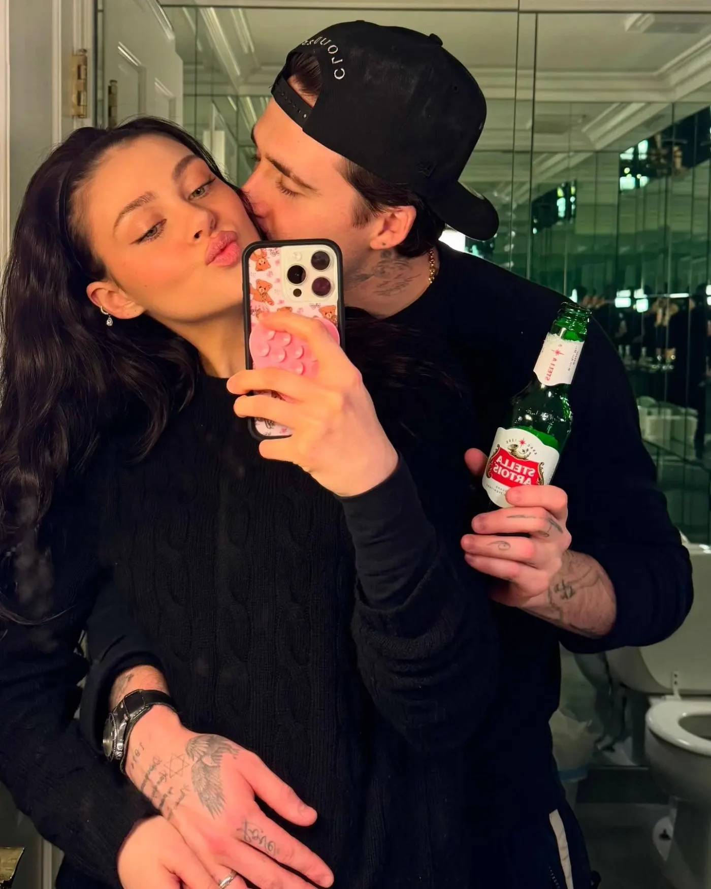 Brooklyn Beckham kissing Nicola Peltz on the cheek while she takes a mirror selfie holding a beer bottle.