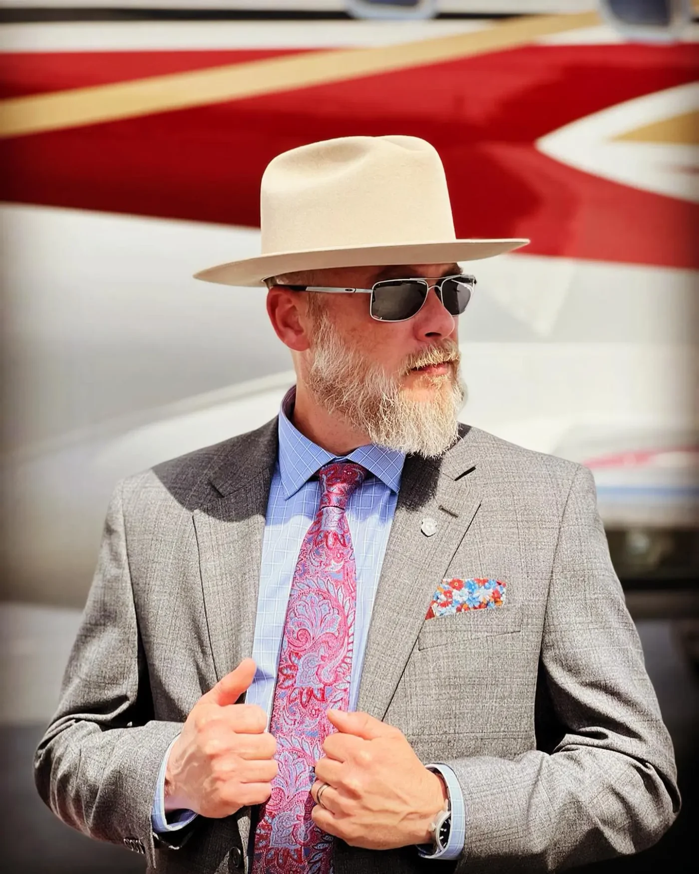 Bryan Seaver wearing a grey suit, fedora, and sunglasses in front of an aircraft.