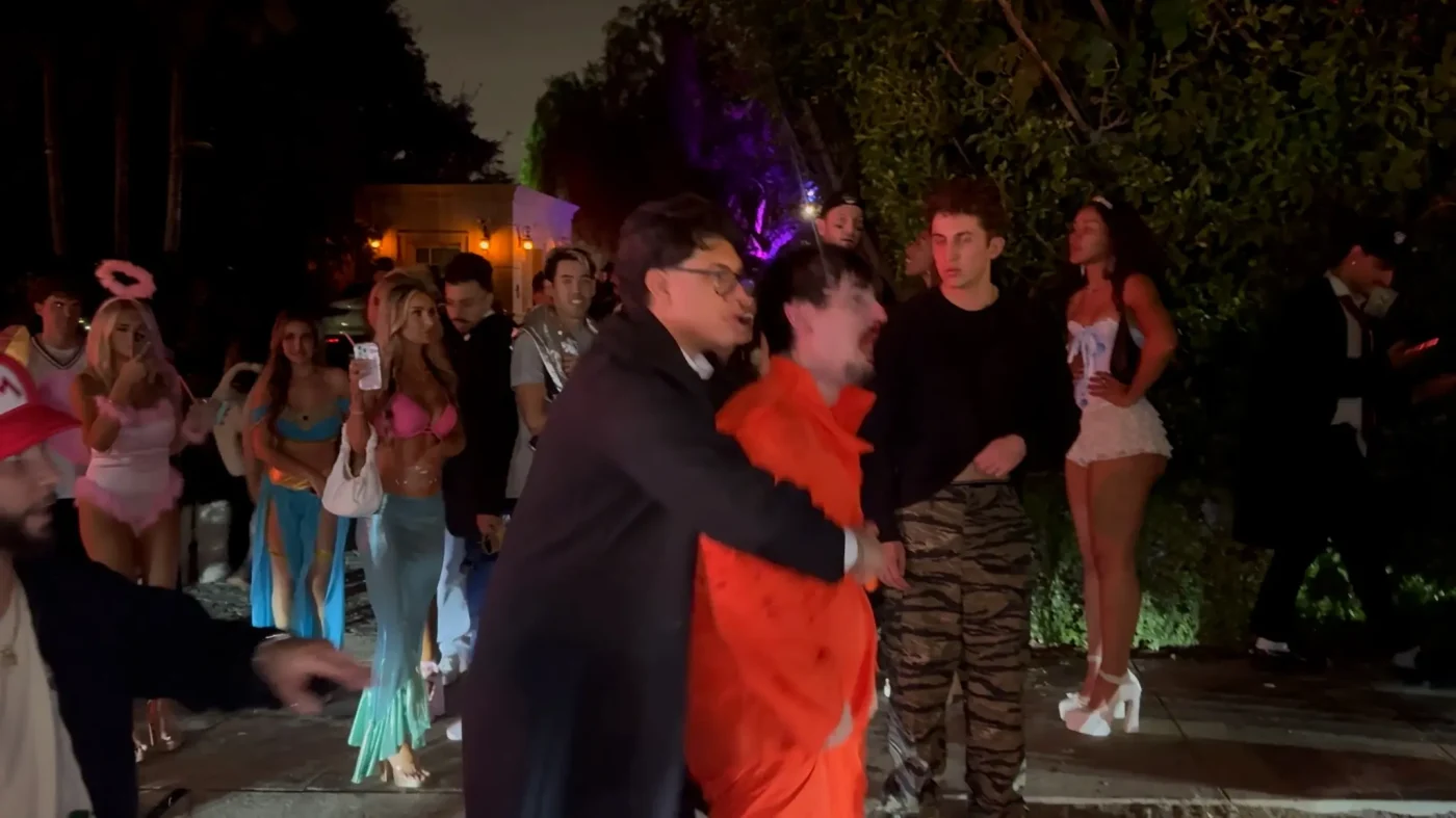 Bryce Hall, dressed in an orange prison jumpsuit, is restrained by a man in a black coat while a crowd of people in Halloween costumes watches.