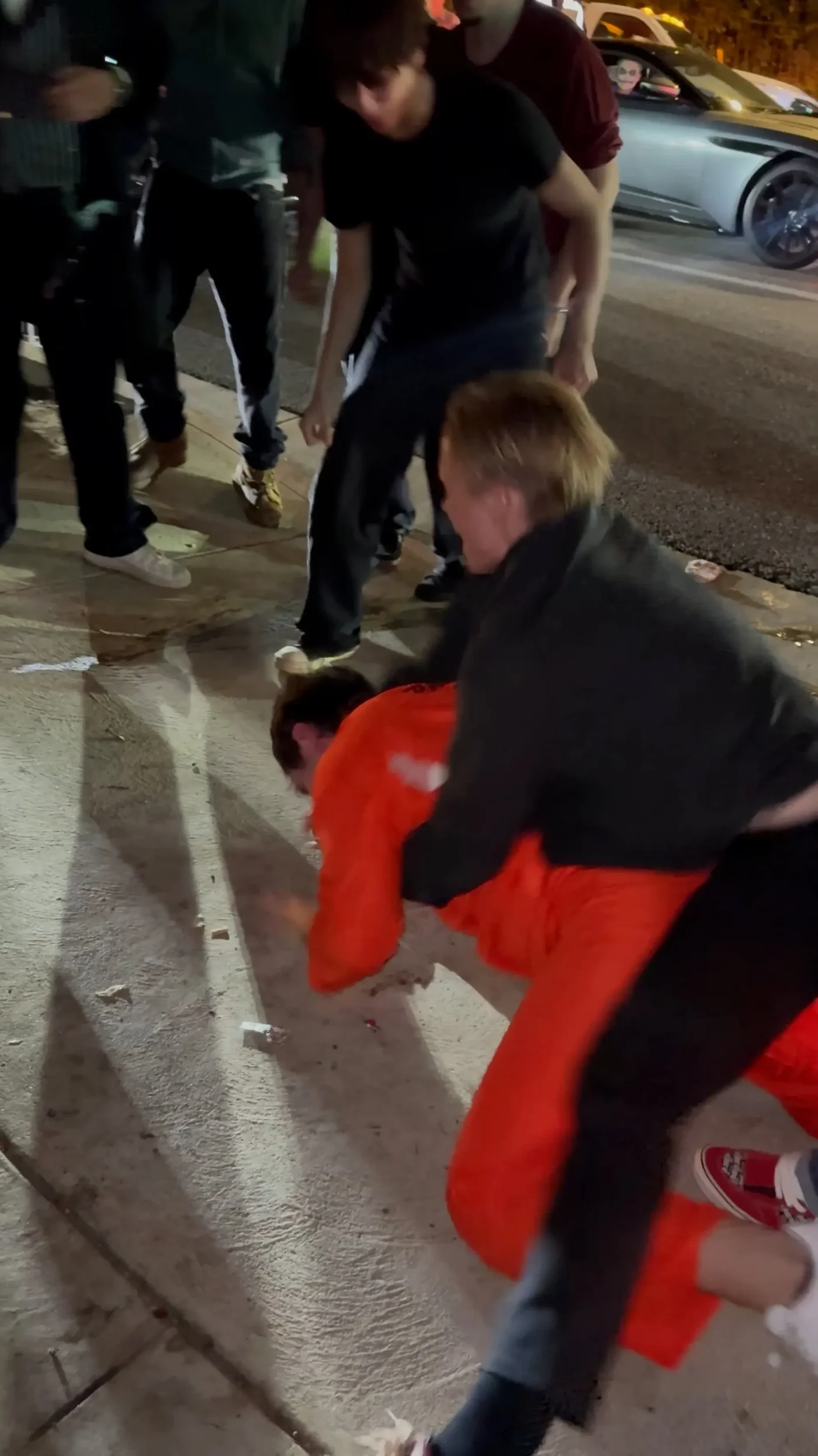 Bryce Hall in an orange prison jumpsuit is wrestled to the ground by a man in a black jacket while onlookers stand nearby on a street.