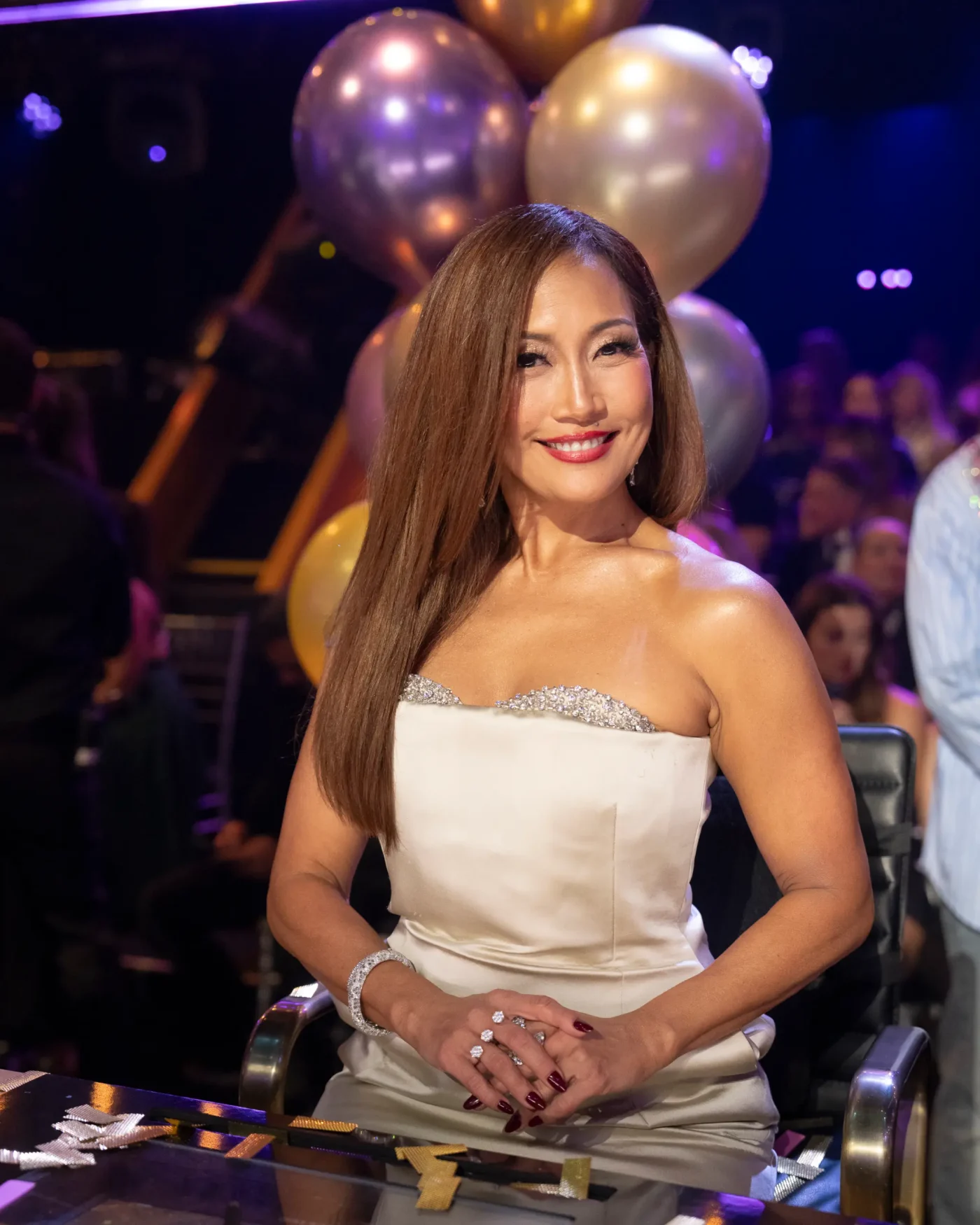 Carrie Ann Inaba smiles in a white strapless dress with silver embellishments, seated with silver and gold balloons in the background.