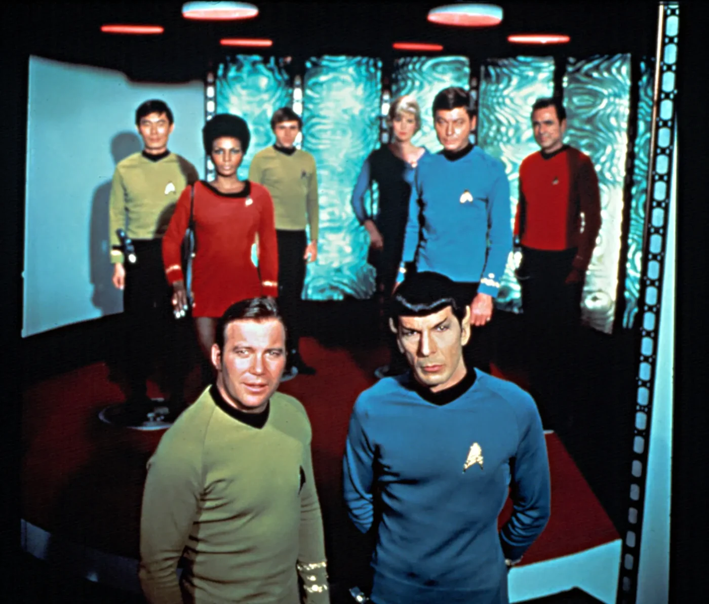 Cast shot of the Star Trek crew on the transporter room set, featuring William Shatner as Captain Kirk and Leonard Nimoy as Spock in the foreground.