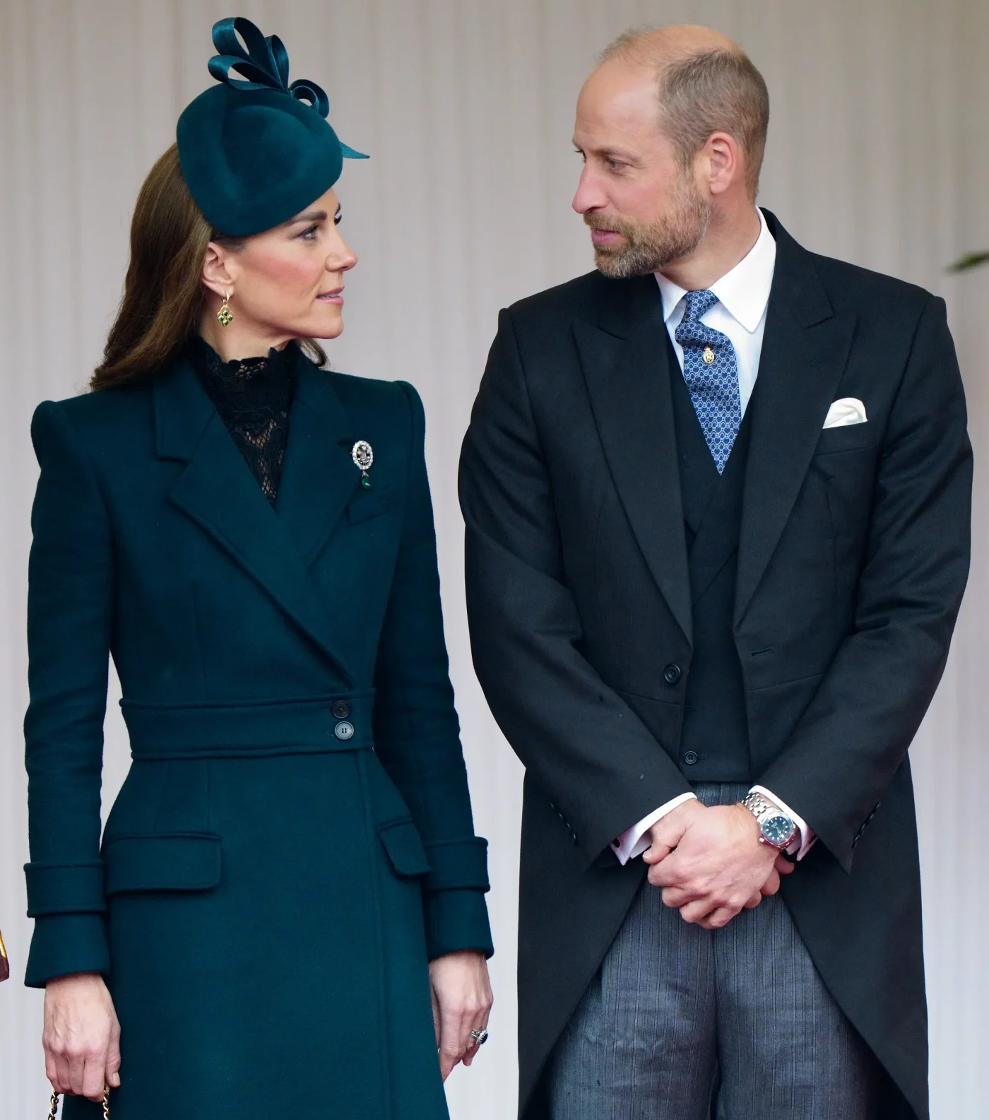 Catherine, Princess of Wales, wearing a dark green coat and matching hat, and Prince William, Prince of Wales, in a suit, look at each other.
