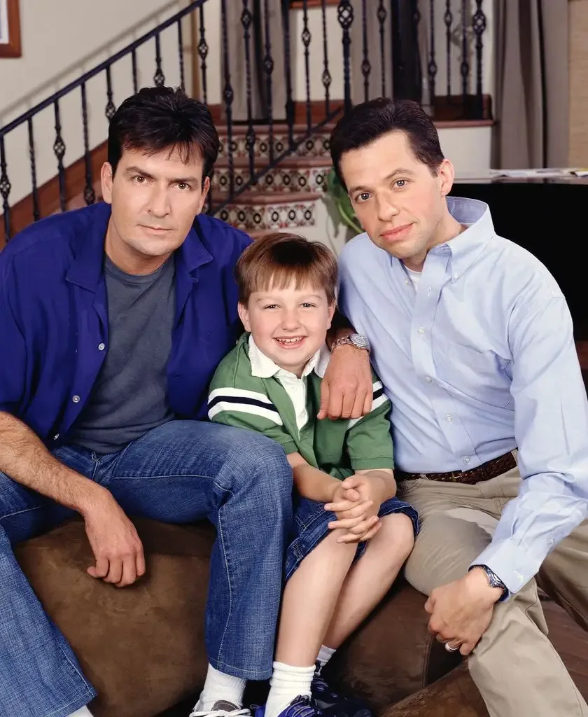 Charlie Sheen, Angus T. Jones and Jon Cryer pose together on a couch in front of a staircase for a Two and a Half Men cast portrait.