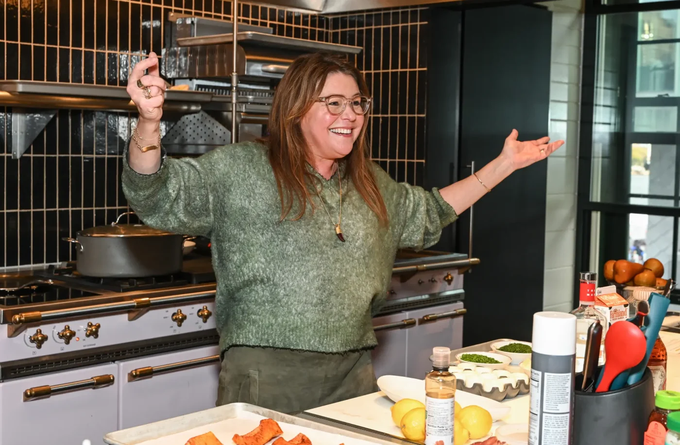 Chef Rachael Ray stands in a kitchen with her arms raised, smiling during a cooking event.