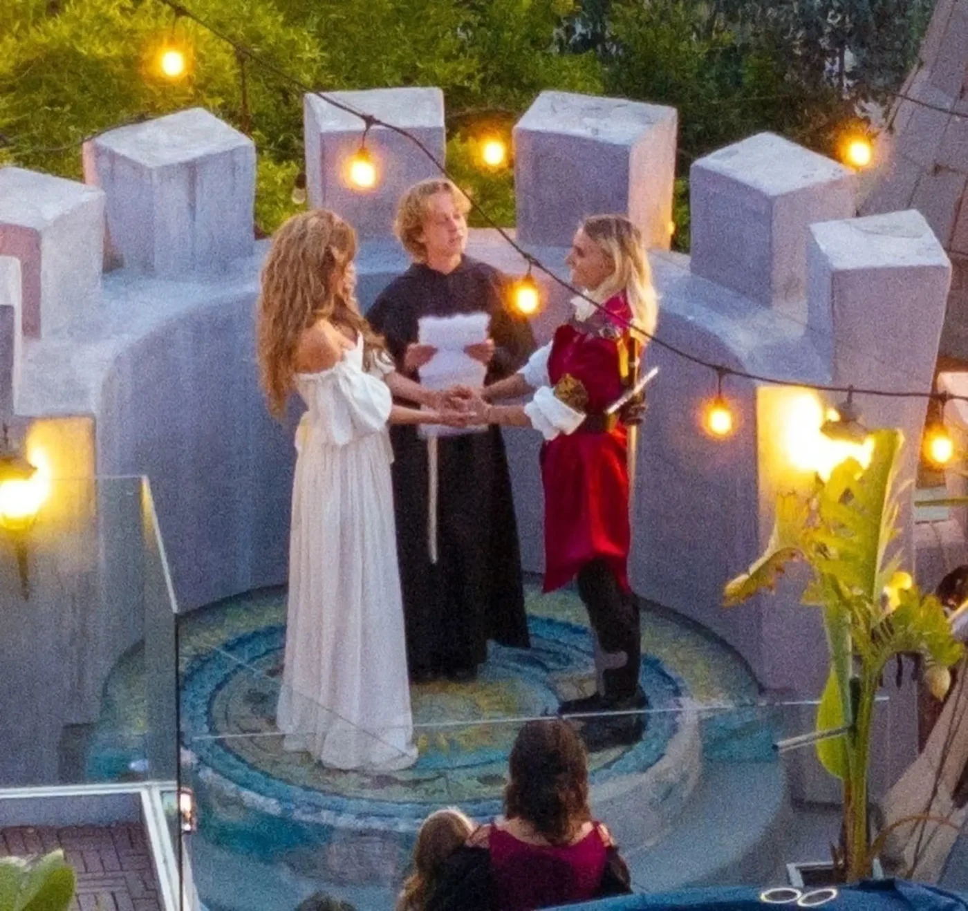 Chrishell Stause in a white gown and G Flip in a red prince-like outfit holding hands during a wedding ceremony on a castle turret.