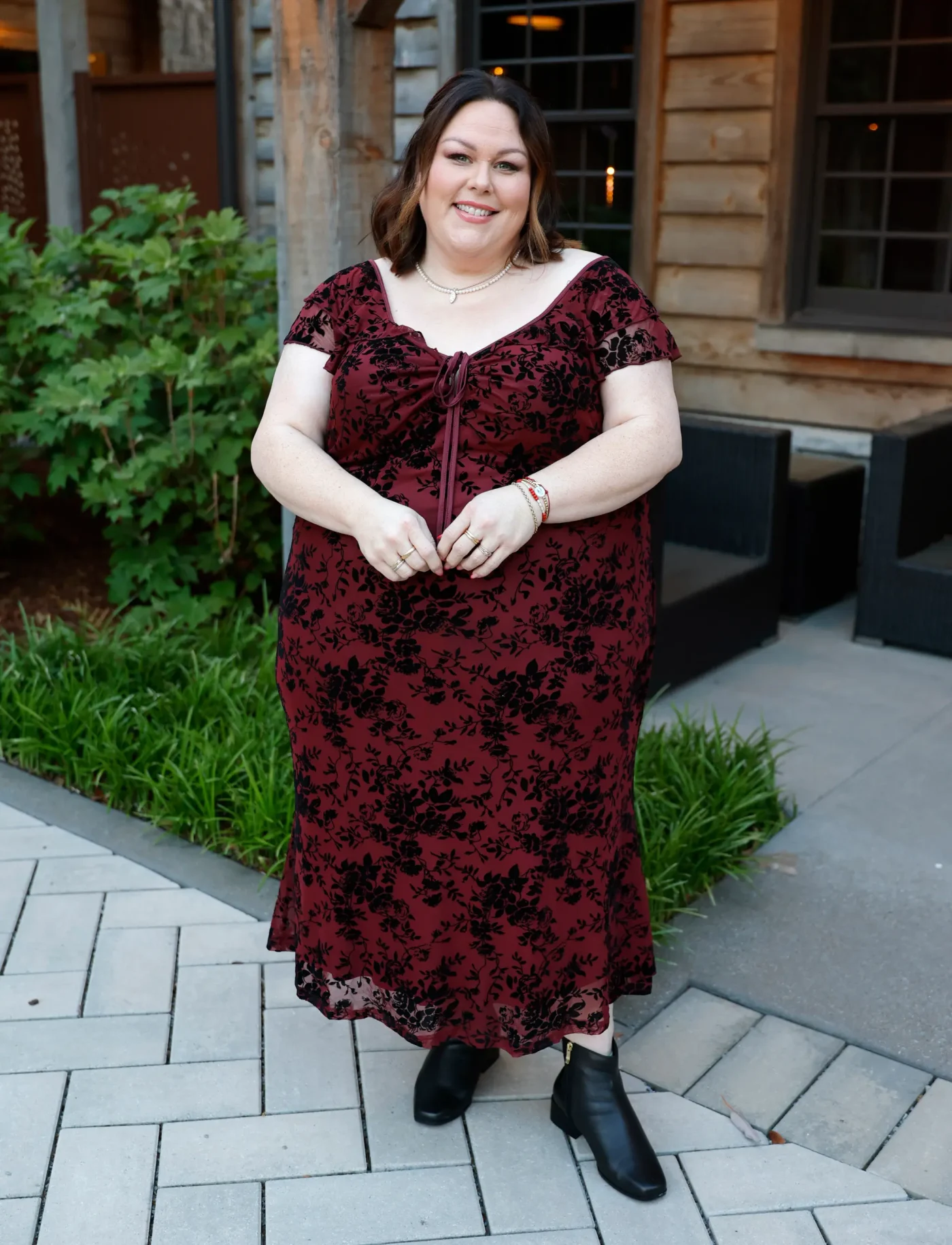 Chrissy Metz poses for photos in a red and black floral dress, pearls, and black boots.