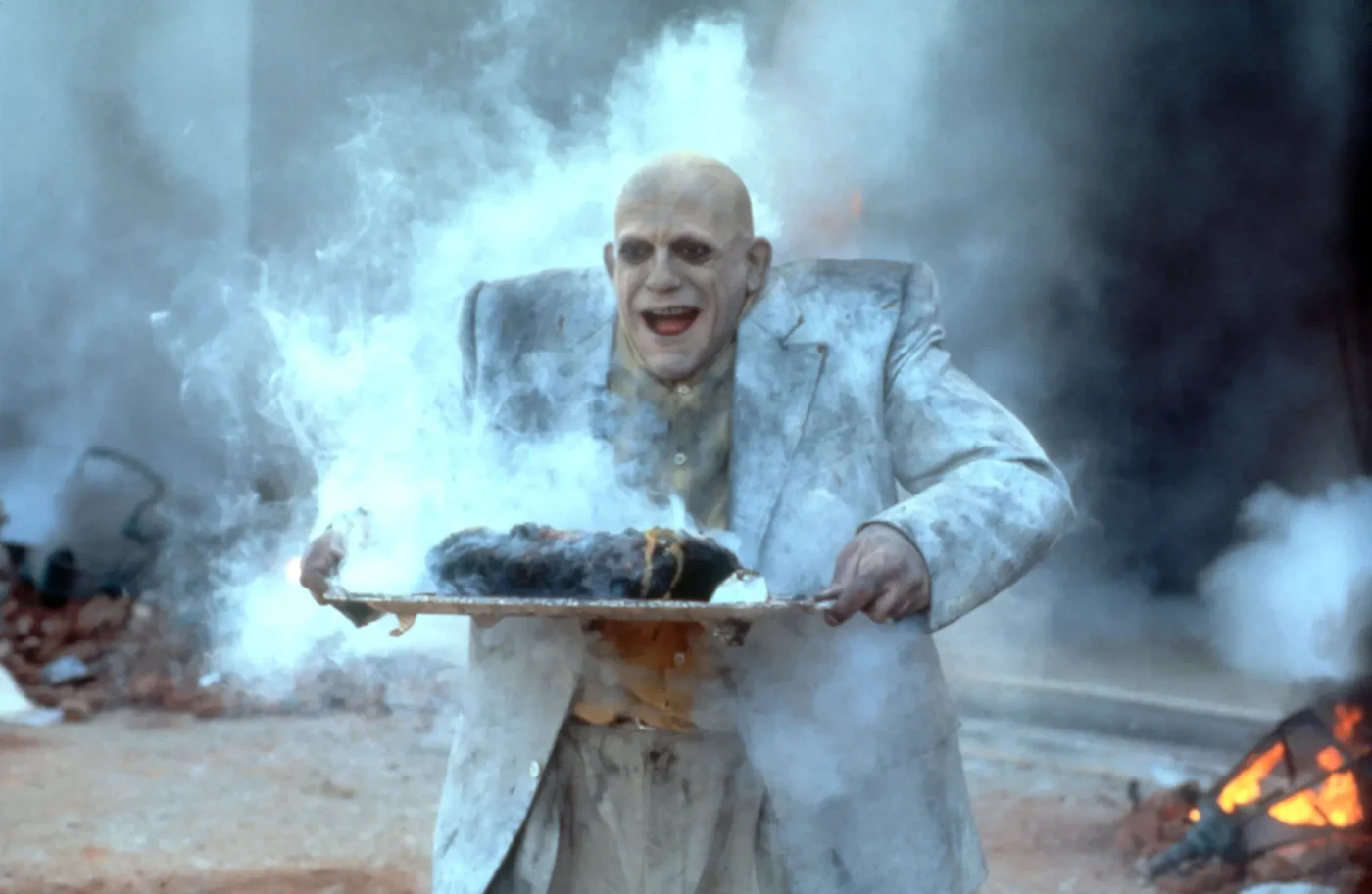 Christopher Lloyd as Uncle Fester holding a smoking platter amidst smoke and debris.