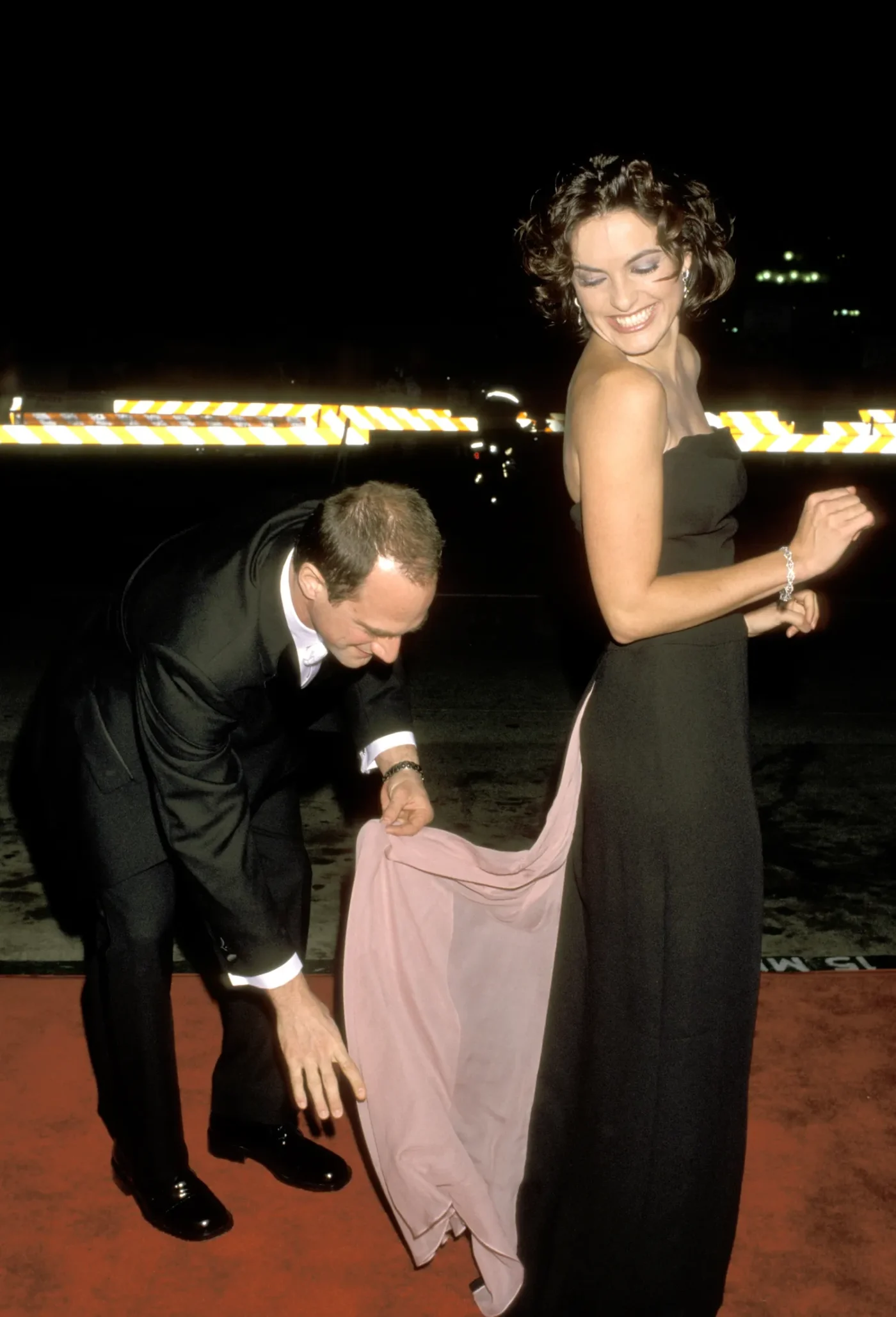 Christopher Meloni adjusting a train of a womans dress on a red carpet