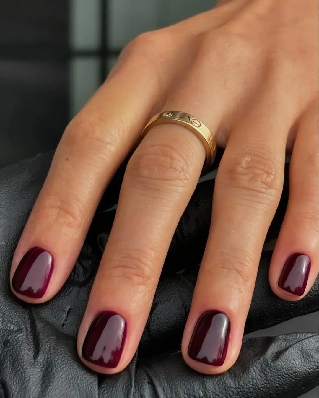 Closeup of a hand wearing a gold ring resting on a textured surface with dark red polished nails