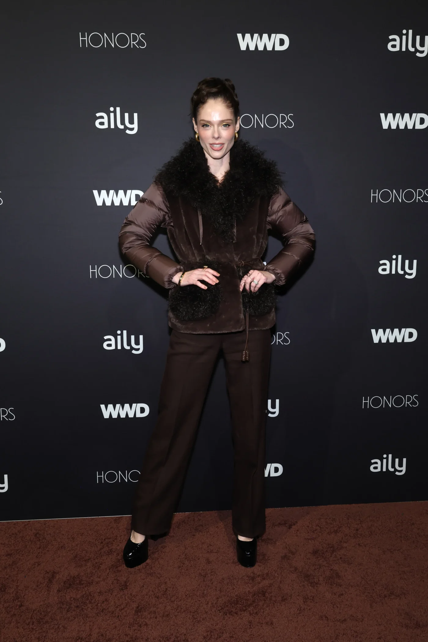 Coco Rocha poses on the red carpet at the WWD Honors in a brown puffy jacket with a fur collar and matching trousers.