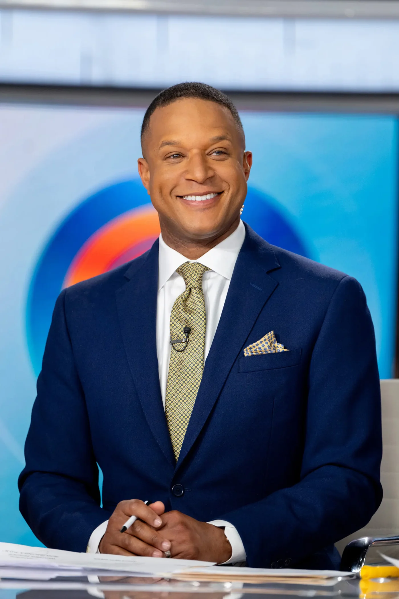 Craig Melvin smiling in a blue suit and yellow patterned tie on the set of the TODAY show.