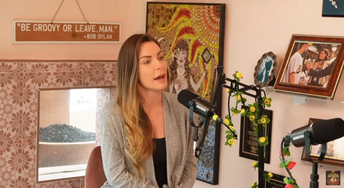 Crystal Hefner sitting in front of microphones in a room with artwork and a sign reading