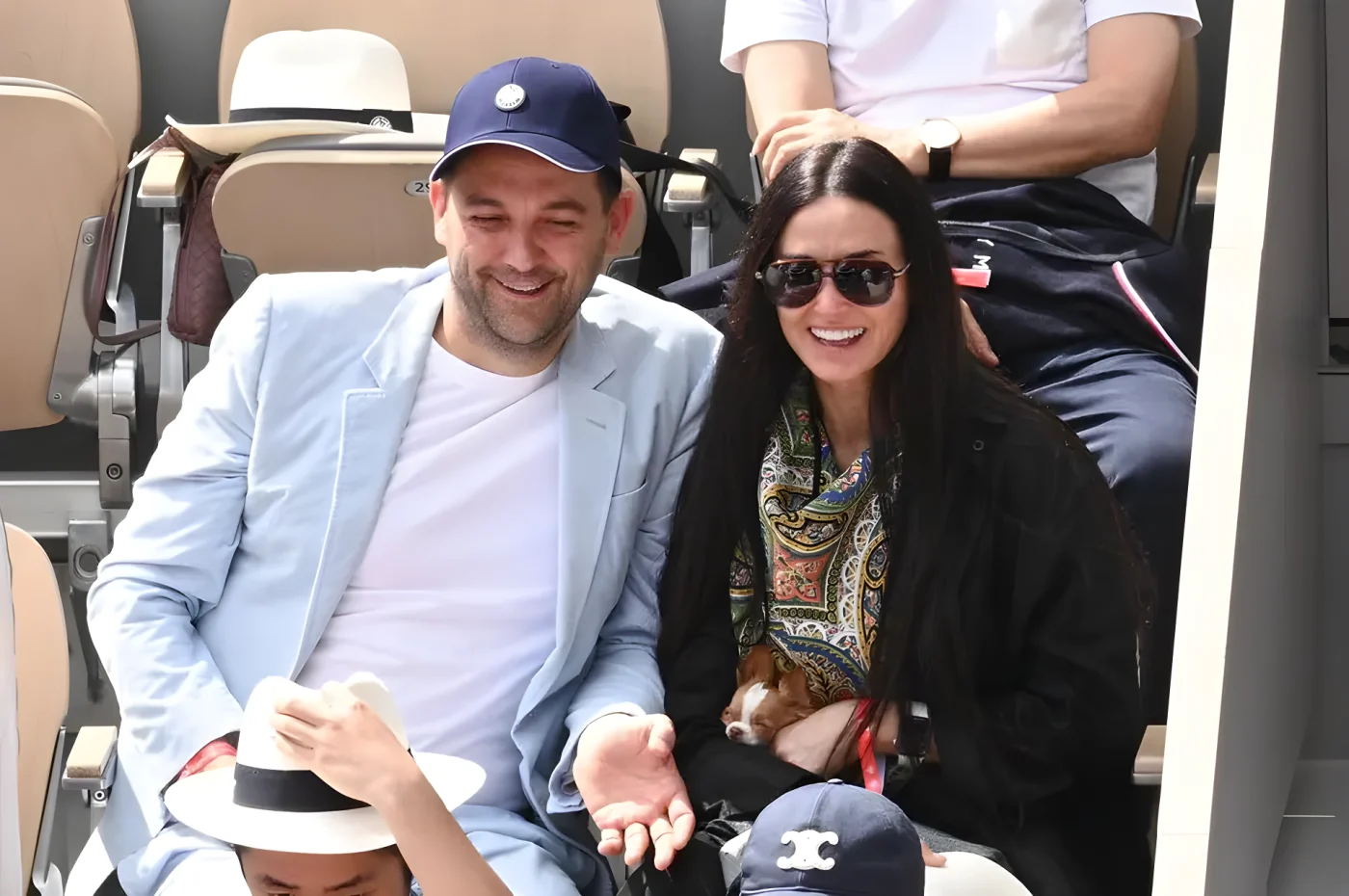 Daniel Humm and Demi Moore sit together smiling in the stands at Roland Garros, with Moore holding a small chihuahua in her arms.