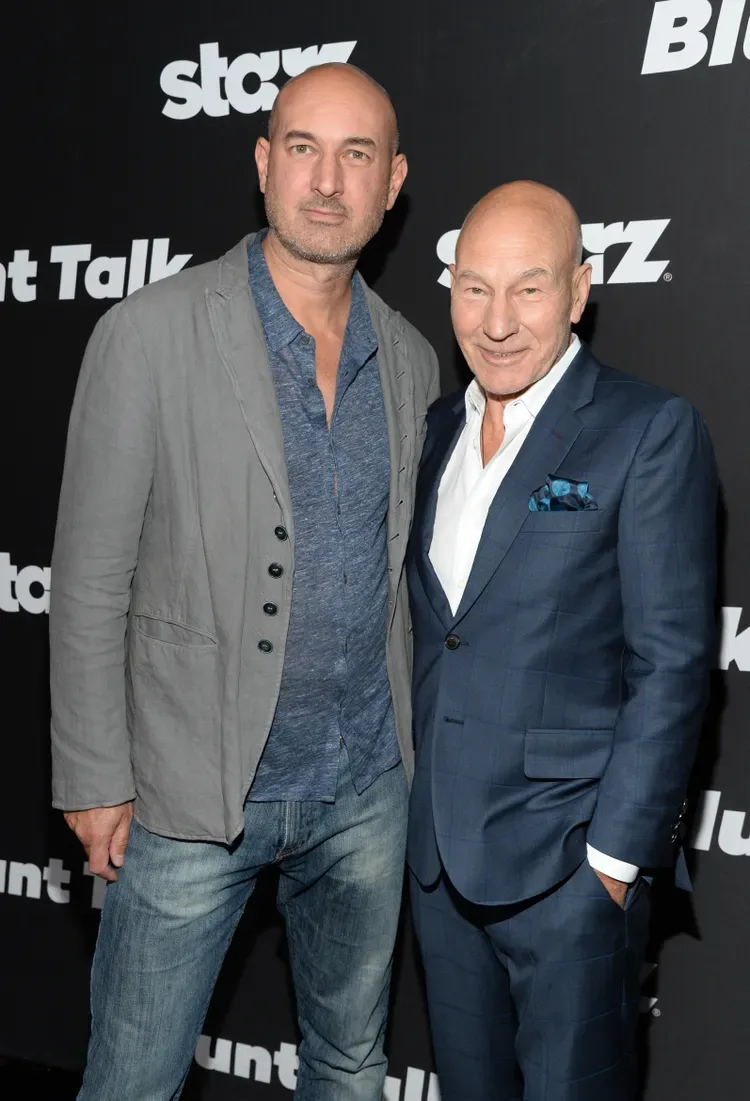 Daniel Stewart and Patrick Stewart pose side by side on the red carpet. Daniel is wearing a gray suit jacket over a heather blue button-up and light-wash jeans. Stewart is wearing a dark blue suit over a white dress shirt and a vibrant blue pocket square.