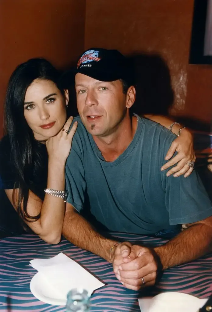 Demi Moore rests her chin on her hand while Bruce Willis, wearing a Planet Hollywood Paris cap, claps his hands in front of him at a restaurant table.