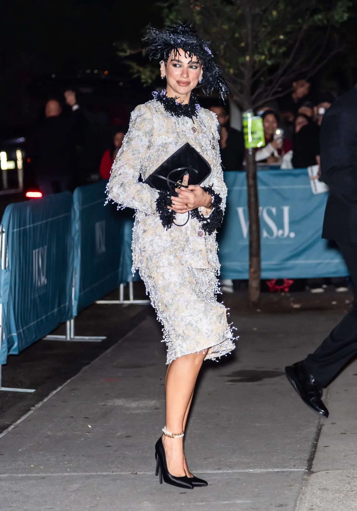 Dua Lipa arriving at the WSJ. Magazine Innovator Awards wearing a textured Chanel skirt suit with black accents, a matching feathered hat, and heels while holding a clutch.