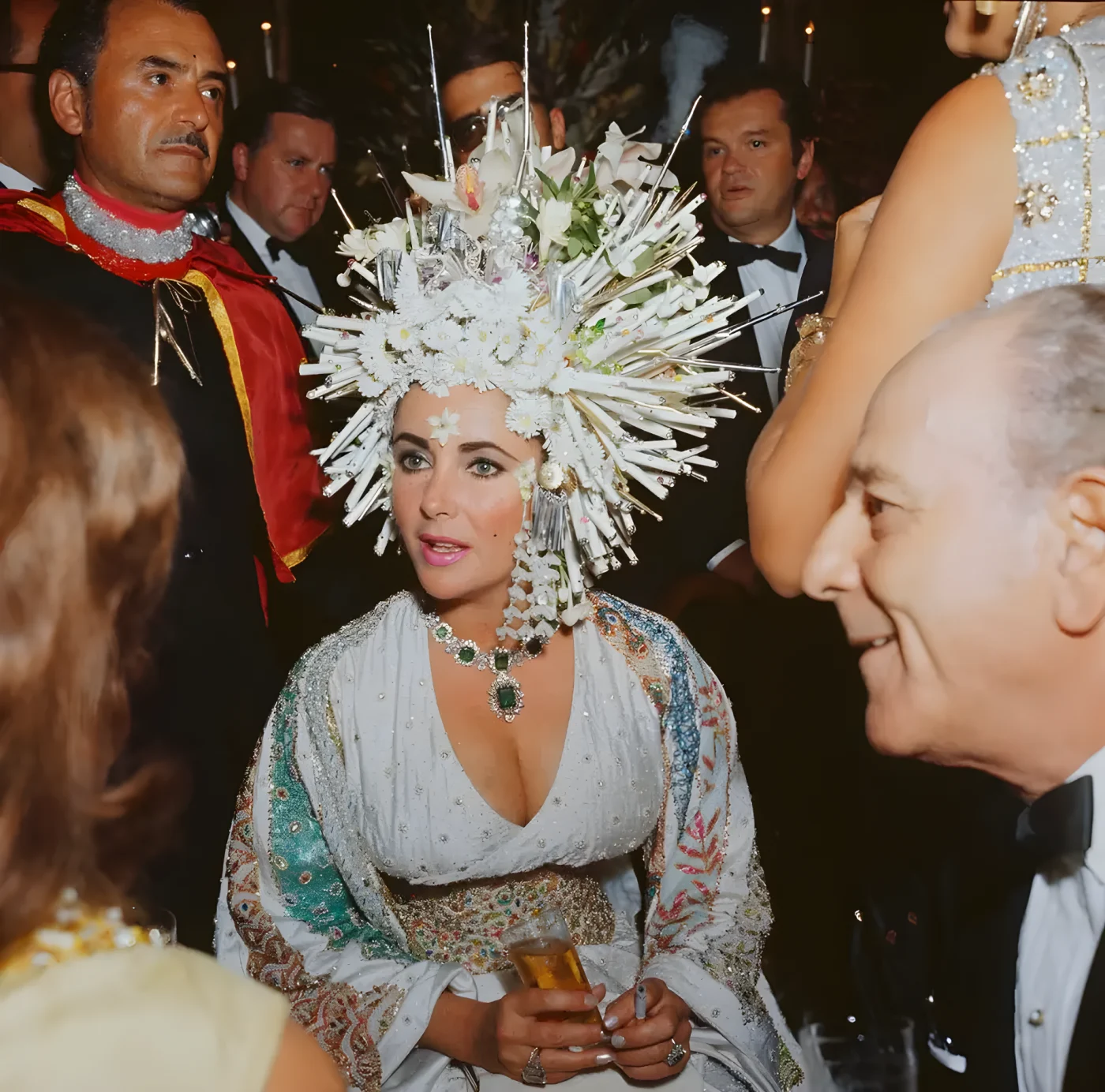 Elizabeth Taylor attends a social function wearing an elaborate headdress of pearls and fake flowers, a jewelled dress and an emerald necklace.