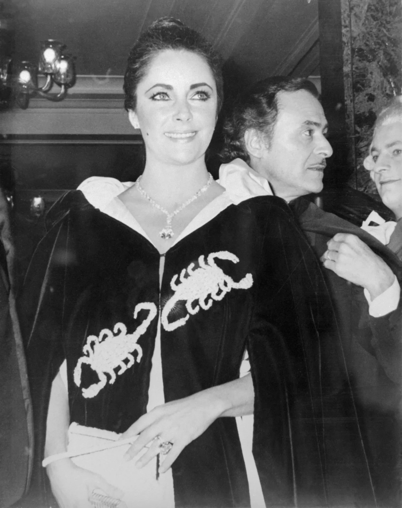 Elizabeth Taylor wearing a diamond necklace and a cape with scorpion appliqués, alongside Richard Burton.