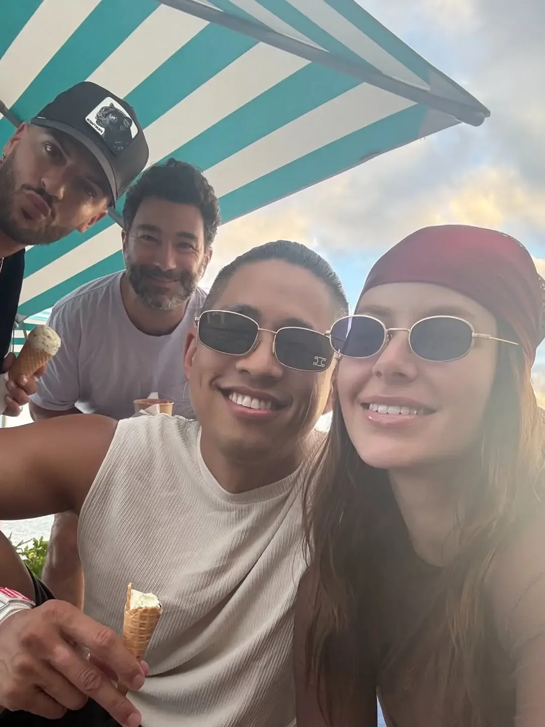 Four people taking a selfie outdoors under a striped umbrella, holding ice cream cones.
