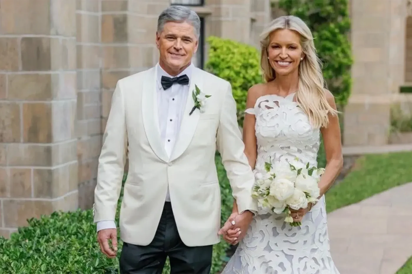 Fox News stars Sean Hannity and Ainsley Earhardt holding hands and smiling in their wedding attire.