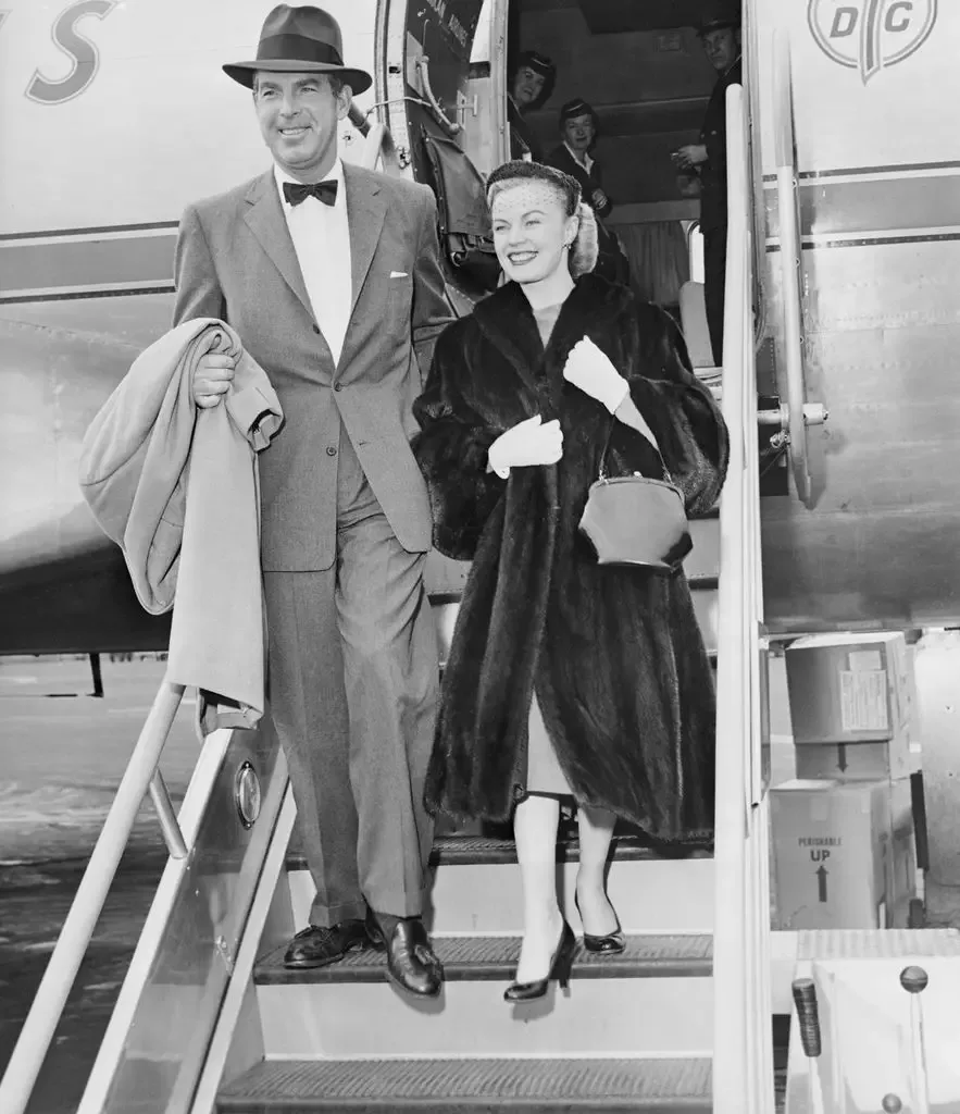 Fred MacMurray in a suit and bow tie and June Haver in a fur coat descend aircraft steps, both smiling, with airline crew visible behind them.