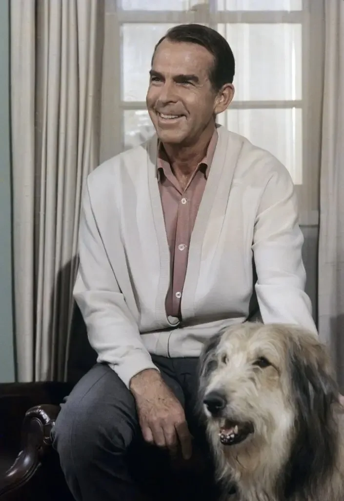Fred MacMurray, smiling in a cream cardigan, sits with the shaggy dog Tramp beside him.