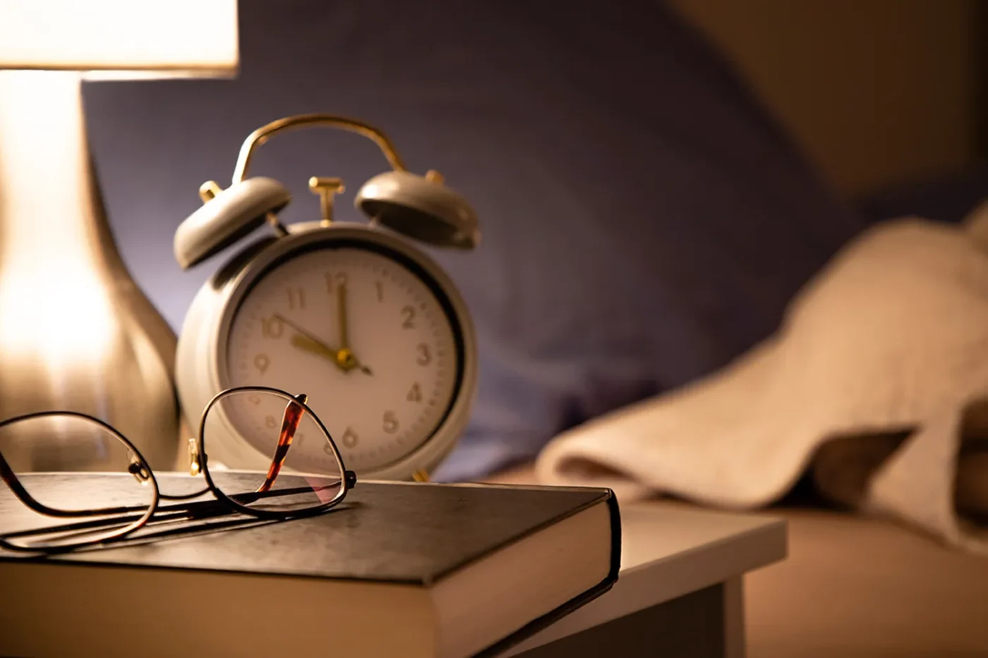Front view of a reading book and an alarm clock on a nightstand with an unmade and defocused bed background at night