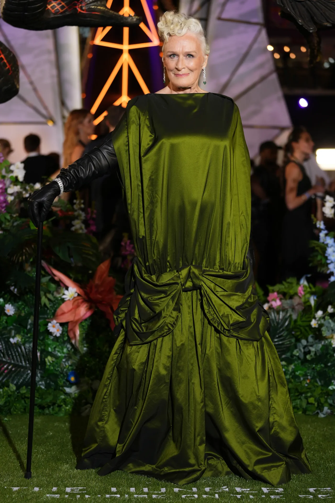 Glenn Close poses in a green dress and long black gloves, holding a cane on a green carpet.