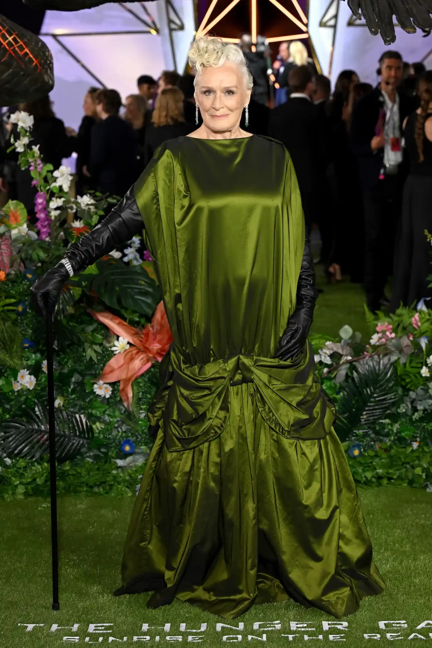 Glenn Close poses in a green satin evening gown with a boat-neckline and emerald drop earrings. She styled the look with lengthy black leather gloves and a matching cane. Her white hair is styled in a pile of curls atop her head.