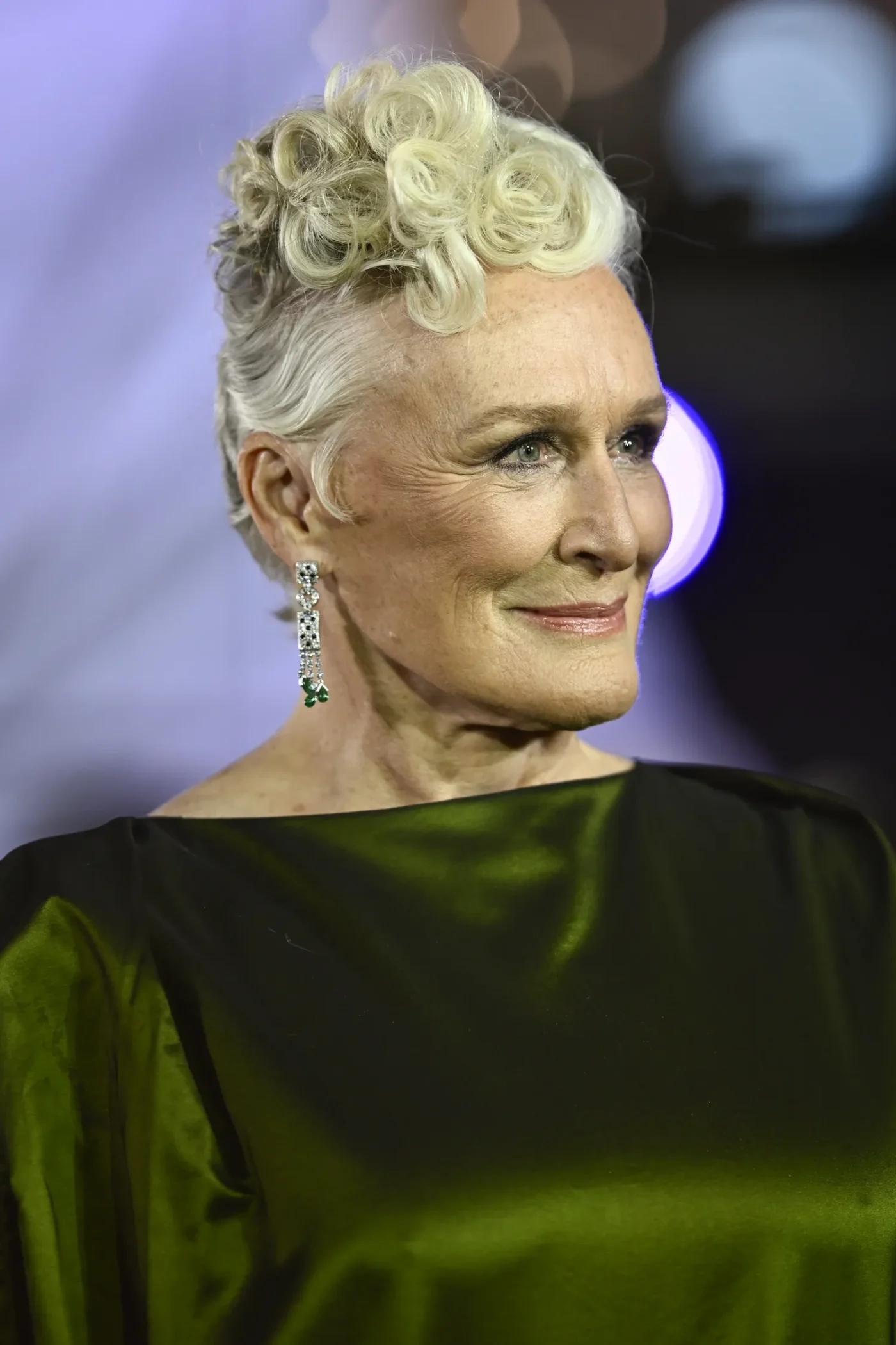 Glenn Close smiles as she looks off into the distance. She is wearing a green satin evening gown with a boat-neckline and emerald drop earrings. Her white hair is styled in a pile of curls atop her head.