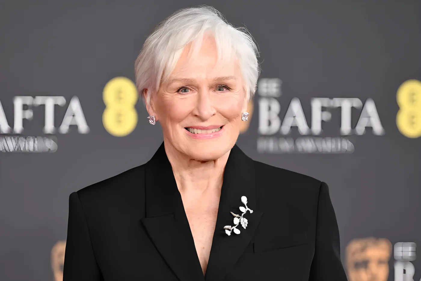 Glenn Close smiles at the camera as she poses in a black blazer with a silver foliage-inspired brooch on her lapel. She is wearing large diamond earrings and her white hair is styled in a soft pixie cut.