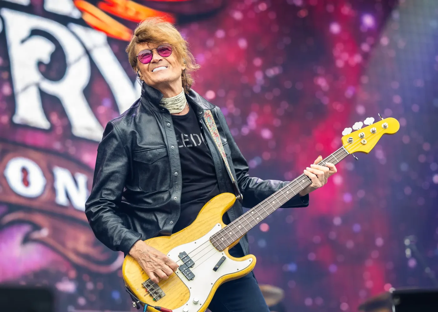 Glenn Hughes of Black Country Communion performing live on stage, wearing sunglasses and a leather jacket while playing a bass guitar.