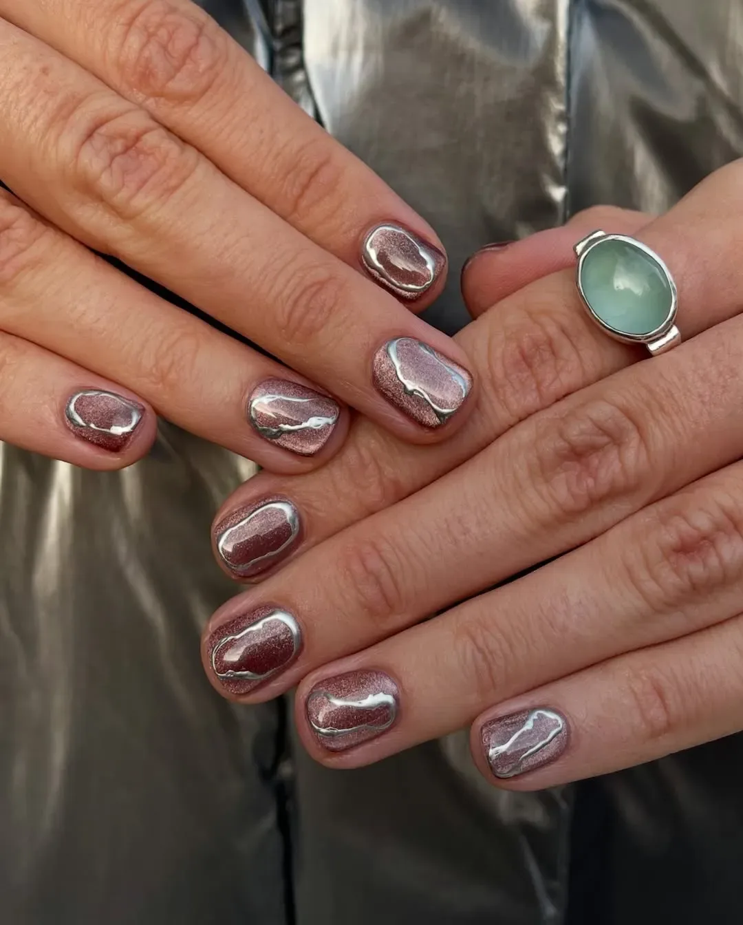 Hands with metallic nail polish and a jade ring on one finger against a metallic background