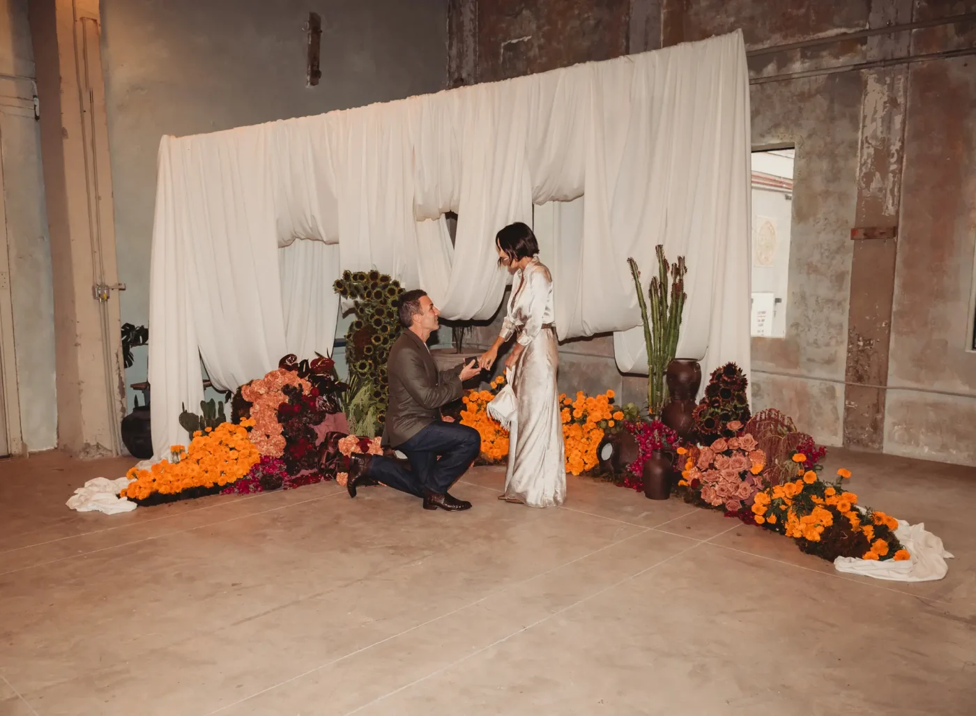 Harvey Spevak proposes to Bianca Peters, who is visibly emotional, in front of a white draped backdrop decorated with orange flowers and cacti.